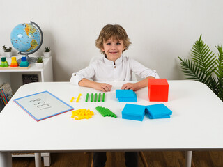 A young boy with a cheerful expression is sitting at a white table, learning about place value and counting using colorful base ten blocks. 