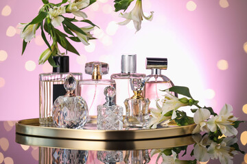 Many different perfume bottles and flowers on mirror surface against pink background with blurred...