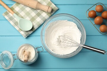 Whisk, whipped cream in bowl, eggs and flour on light blue wooden table, flat lay