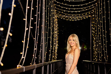 Elegant woman poses under glowing fairy lights on a bridge during a nighttime event near a serene setting