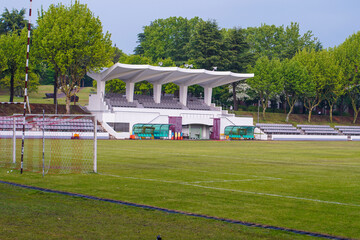 Soccer stadium with green field, white goalposts, and covered seating. Surrounded by trees under an overcast sky.