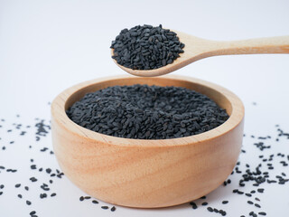Wooden spoon lifting black sesame seeds from a wooden bowl with some seeds spilled on the table.