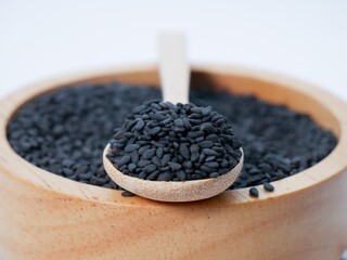 Close-up of a wooden spoon resting in a wooden bowl filled with black sesame seeds on a white background.