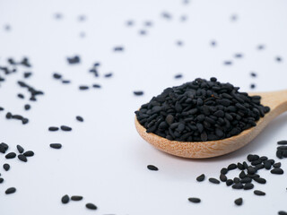 Black sesame seeds in a wooden spoon with scattered seeds around on a clean white background.