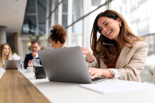 Businesswoman planning over phone call while researching on laptop in board room with colleagues working in the background