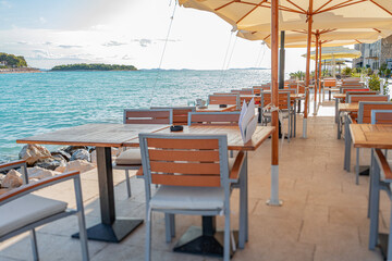 Tranquil seaside restaurant with modern wooden tables and chairs arranged under large umbrellas along a scenic waterfront promenade.