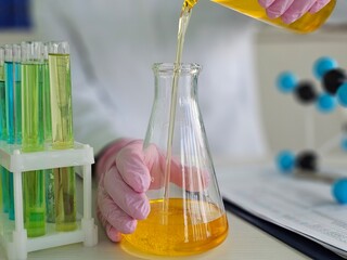 A scientist in a lab coat wears gloves while carefully pouring a bright yellow liquid from a test...