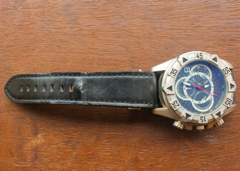 Luxury analog wristwatch with a black leather strap and stainless steel bezel, placed on a wooden surface, showcasing bold numerals and a modern chronograph dial.