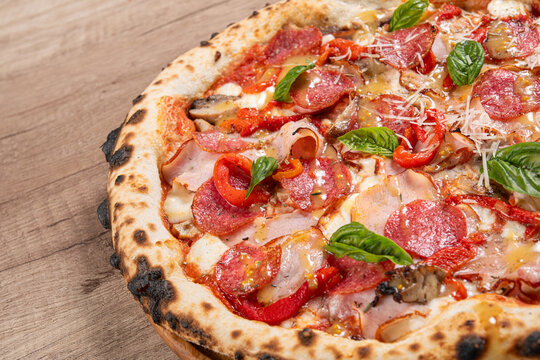 A close-up image of a wood-fired pizza topped with pepperoni, ham, red bell peppers