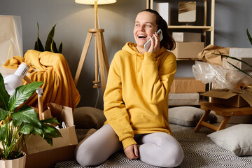 A woman sits on the floor among moving boxes, laughing while talking on her phone after moving into her new home.