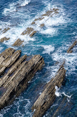 Fototapeta premium Dynamic aerial view of rugged rocky shoreline with turquoise ocean waves splashing against sharp stone ridges, showing natural power and coastal landscape beauty.