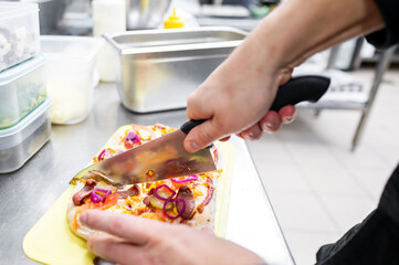 A chef slices a freshly baked pizza with a knife on a stainless steel counter in a professional kitchen. The pizza is topped with cheese, pepperoni, and red onions