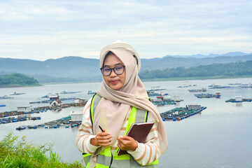 A female fish farm manager wears a safety vest and carries a book to check the work.