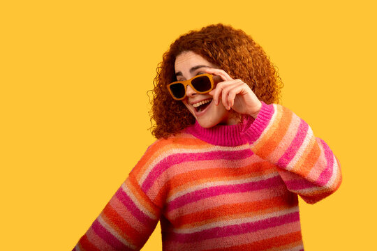 Cheerful redhead woman lowering sunglasses and smiling on yellow background
