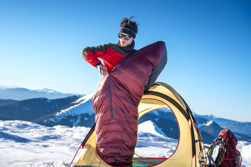Young man standing halfway out of a tent while zipping up a sleeping bag in a snowy mountain landscape. Winter camping scene with outdoor gear and clear blue sky © XArt