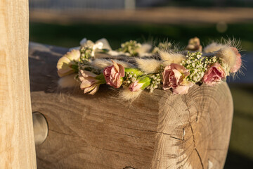 A delicate floral crown made of soft pink roses, baby's breath, and wheat ears rests on a natural wooden log. A symbol of nature, simplicity, and festive tradition.