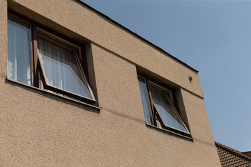 There are two open windows on a beigecolored building that stand out against the backdrop of a clear blue sky above