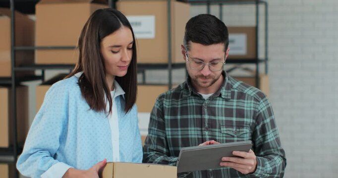 A man sales manager using a tablet computer is talking to an employee holding a cardboard box. A warehouse with parcels ready for shipment in the background.