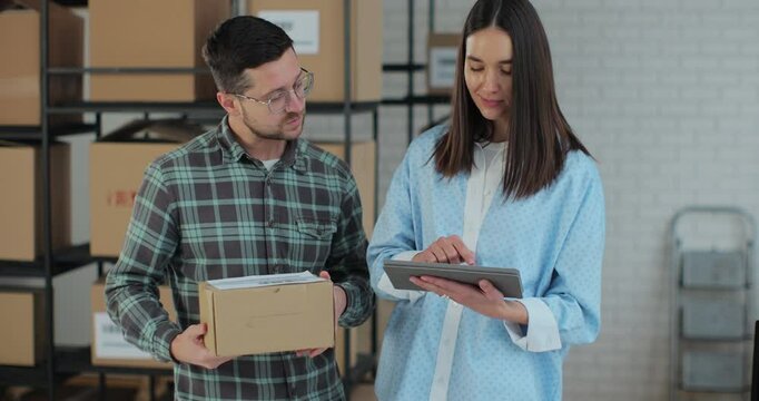 A female sales manager using a tablet computer is talking to a male worker holding a cardboard package. A warehouse with parcels ready for shipment in the background.