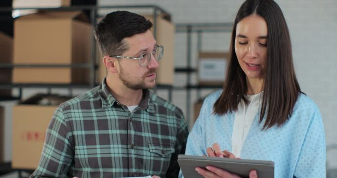 Close-up of a female manager showing information on a tablet computer to an employee with a cardboard box. Stock of parcels with products ready for shipment in the background.