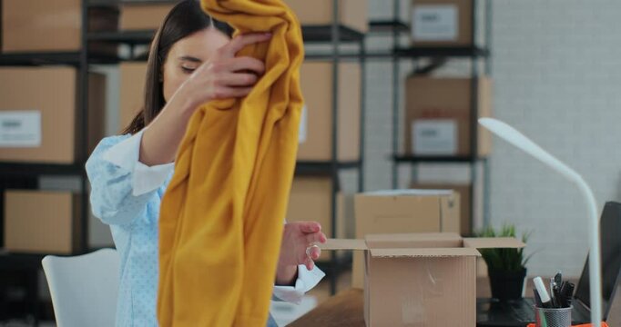 An employee packs a yellow jumper in a room with shelves full of parcels. The woman owner of an online store works on a computer at his desk in a warehouse.