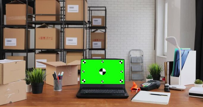 Laptop with green screen chromakey display mockup standing on desktop in warehouse. Cardboard boxes in the background.