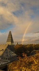 Cityscape with modern glass tower and rainbow after rain, captured at golden hour with autumn foliage in the foreground. © olga
