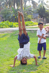 A cheerful schoolboy doing a handstand on the grass while another boy in uniform stands nearby, smiling and watching him