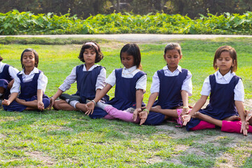 A row of schoolgirls in uniform sit cross-legged on the grass, eyes closed and hands extended forward in a calm and focused meditation session