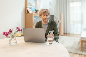 Senior woman using laptop at home while looking at a smartphone screen. Concept of technology, online shopping, digital literacy for seniors and active aging. Technology usage among seniors.