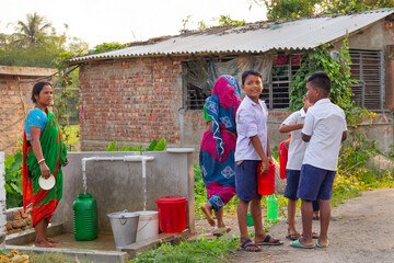 Schoolboys and women gather around a community water point in a village, chatting and waiting their turn to fill containers with water