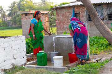 Two women in vibrant sarees fill containers with water at a community tap in a rural village