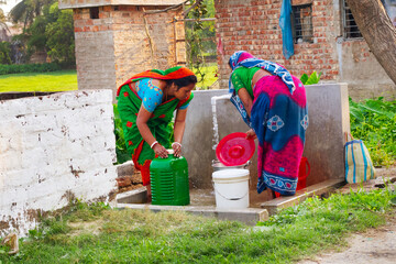 Two women in vibrant sarees fill containers with water at a community tap in a rural village