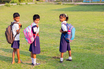 Three schoolchildren with backpacks stand on a grassy field, looking back over their shoulders as they head home after school