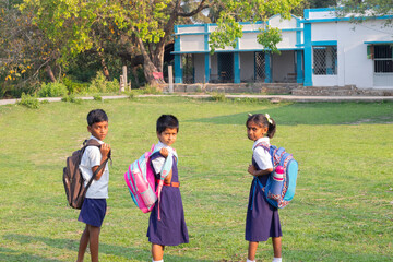 Three schoolchildren with backpacks look back while walking across a grassy school ground, with a small rural school building visible in the background