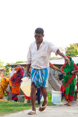 A man in a white shirt and checked lungi walks away from a village water point carrying a yellow container