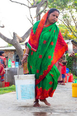 A woman in a green and red saree carrying a heavy white bucket filled with water