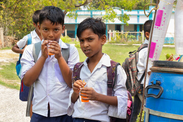 Two schoolboys in uniform drink orange beverages from plastic cups, standing near a refreshment cart outside their school with backpacks