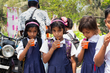 Three schoolgirls in uniform sip orange drinks through straws, standing near a roadside vendor after school on a sunny day