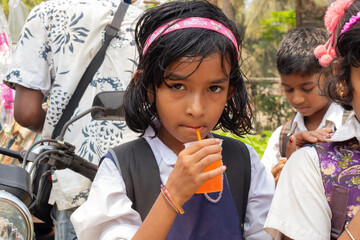 A schoolgirl in uniform with a pink headband drinks an orange beverage