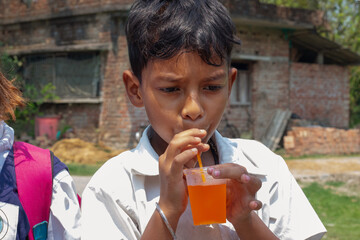A schoolboy in uniform sips an orange drink through a straw, standing outdoors near a rustic brick building after school