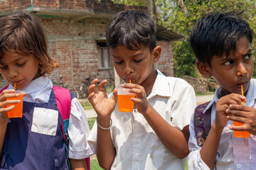Three schoolchildren in uniform enjoy orange drinks under the sun, standing together outside a rustic brick building after class