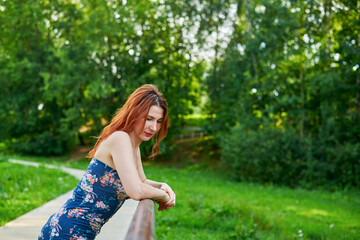 Woman in blue dress leaning on bridge railing in park. Relaxation and rest concept as woman enjoying her time outdoors. Lush green surroundings. Girl has beautiful body.