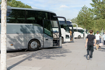 Travelers walking beside parked buses near a city park on a sunny afternoon
