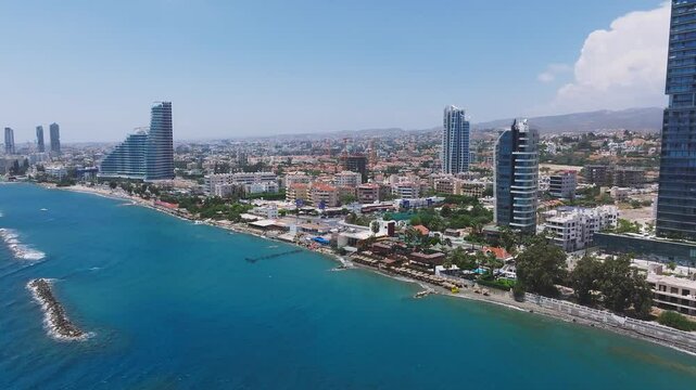 Sweeping drone view of Limassol, Cyprus, featuring a tall blue skyscraper, urban buildings, coastline, piers, greenery, and distant hills.