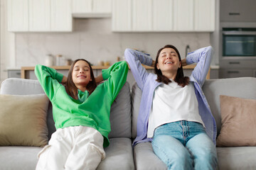 Calm and relaxed mom and teenage daughter chilling on couch hands over head, resting with eyes...