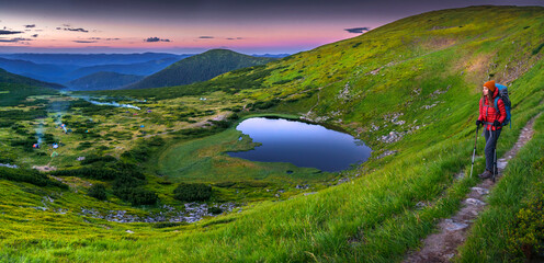 Panoramic view of a female hiker with backpack and trekking poles standing on a mountain trail at...