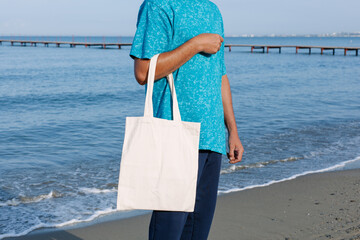 Man with black, tan skin holding in hand white eco cotton textile bag. Blue water of sea or ocean...
