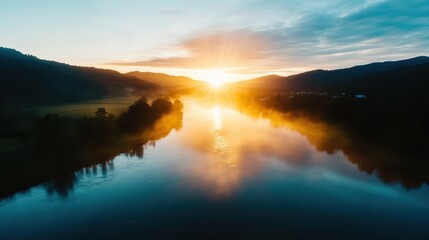 A breathtaking sunrise casts golden hues over a tranquil river, surrounded by mountains and lush greenery, creating a peaceful natural scene at dawn.