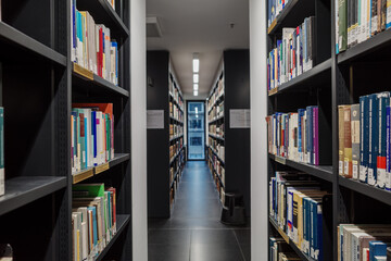 A perspective shot down a long, narrow aisle in a modern library, flanked on both sides by tall bookshelves filled with various books.  © Peeradontax